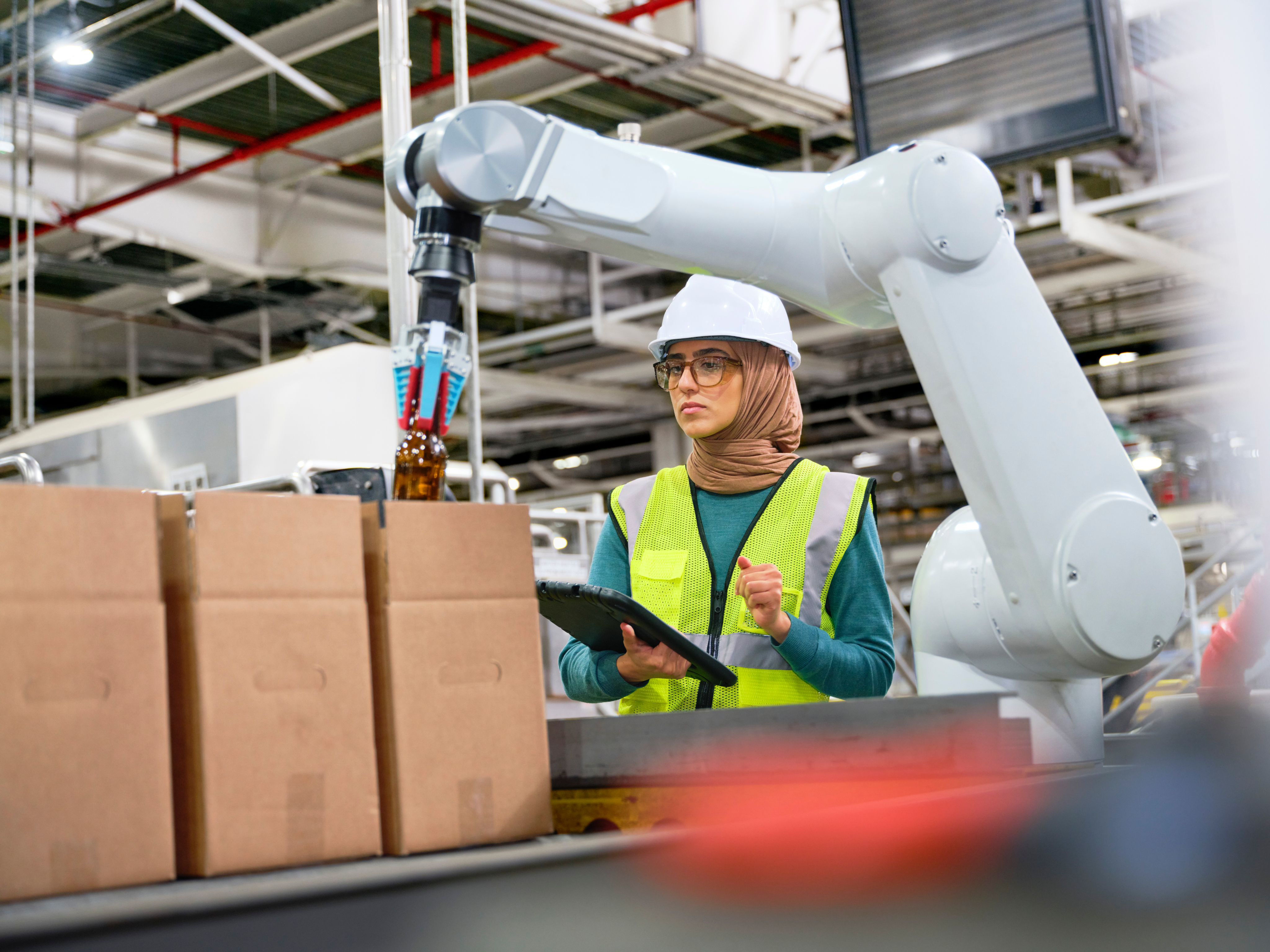 A female worker wearing safety gear using a tablet to monitor a robotic arm on a factory production line.